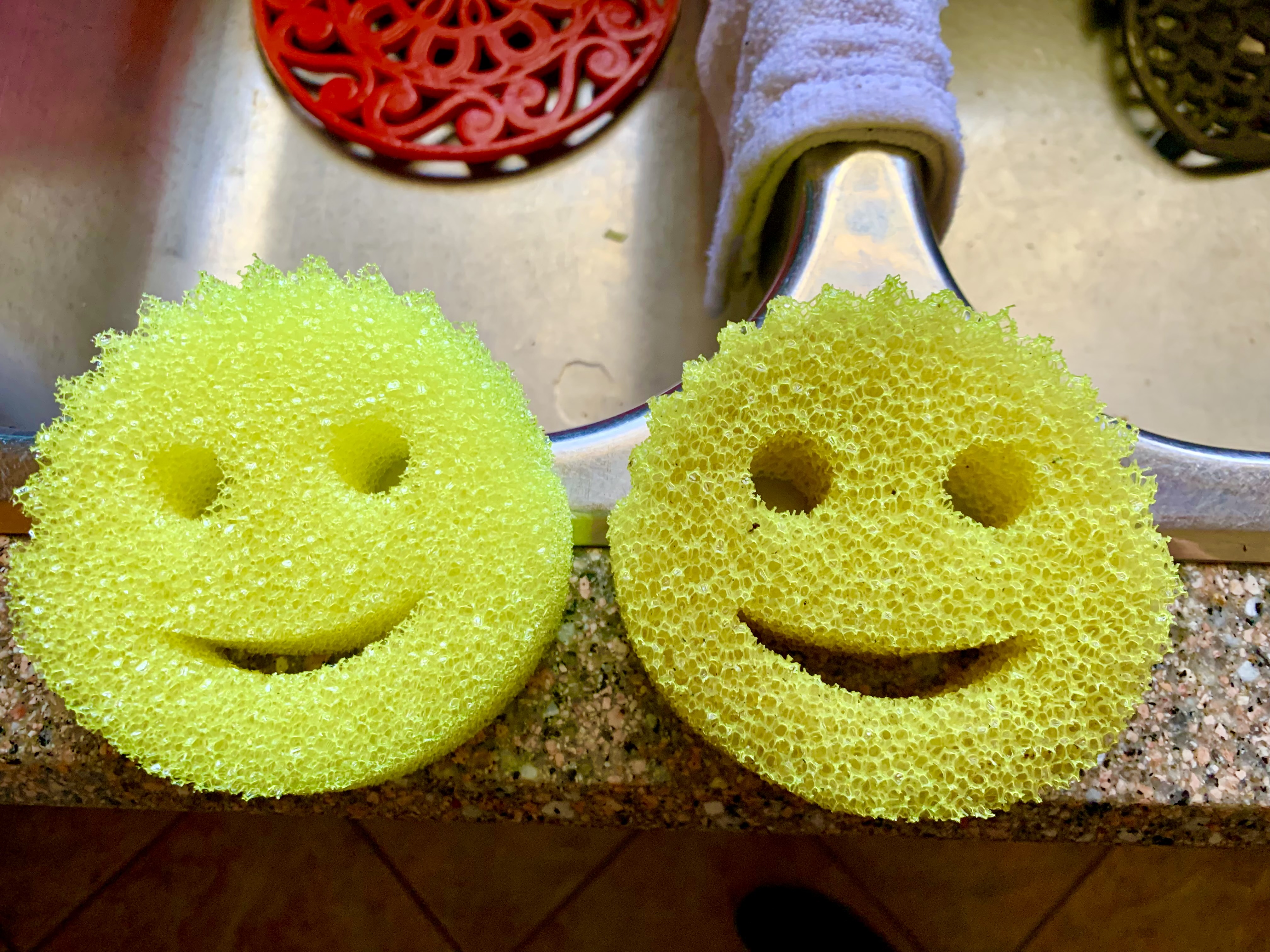 Two yellow smile-shaped Scrub Daddy sponges beside a sink; one visibly more worn.